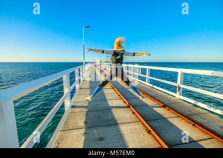 Carefree young sporty woman jumping at Busselton jetty in Busselton, Western Australia. Happy female jumper over iconic wooden pier in WA. Australia travel and freedom concept. Copy space. Stock Photo