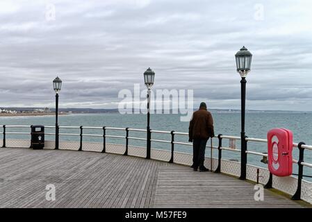 Rear view of a single old gentleman with walking stick looking out to sea on the end of Worthing pier Sussex UK on a dull winters day
