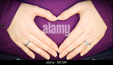 pregnant caucasian woman with hands in heart shape over expectant belly wearing magenta top. beautiful hands with wedding ring