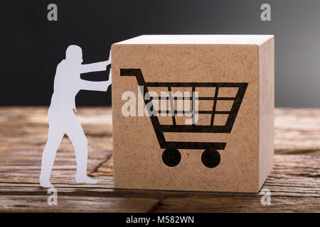 Closeup of paper man pushing wooden block with shopping cart on table against gray background Stock Photo