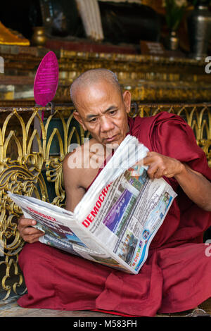 A monk in a maroon-colored robe reading the latest newspaper. Yangon, Myanmar Stock Photo