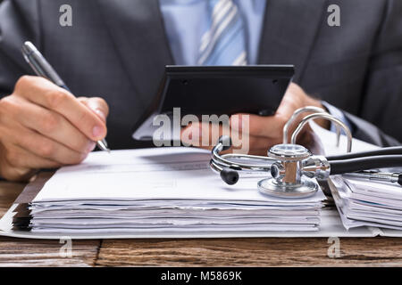 Midsection of businessman calculating invoice with stethoscope on documents at table Stock Photo