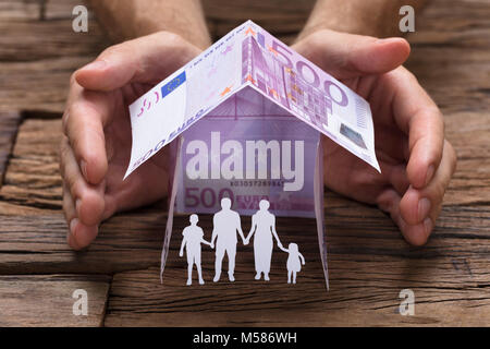 Businessman's hands covering paper family under house made from 500 euro papernotes on wooden table Stock Photo