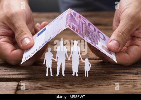 Hands covering paper family under house roof made from 500 euro papernote on wooden table Stock Photo
