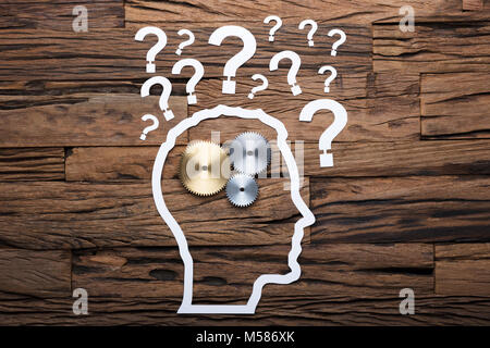 Paper businessman's head outline with metallic gears and question marks on wooden table Stock Photo