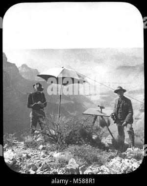 Grand Canyon Matthes Survey of . BLACK AND WHITE GLASS LANTERN SLIDE, LABEL READING 'PLANE TABLE STATION ON CAPE ROYAL, KAIBAB PLATEAU, GRAND CANYON, ARIZ. F.E.M.' TWO MEN SURVEYING ONE WRITING IN A BOOK, THE OTHER LOOKING AT THE CAMERA, THERE IS A LARGE UMBRELLA PRESENT BEHIND THE MEN.  USGS LABEL ATTACHED. CIRCA 1902. WITH A GROUP OF SLIDES FROM THE MATTHES SURVEY EXPEDITION IN THE GRAND CANYON - CIRCA 1902 Stock Photo