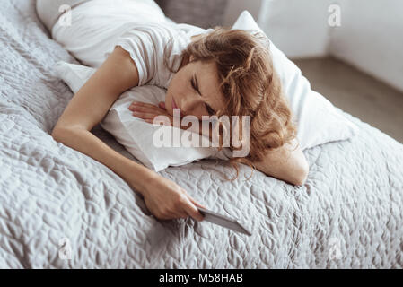 Sad brunette lying on bed with smartphone in her hand Stock Photo