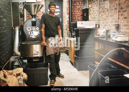 African worker standing in a coffee roasting room next to a coffee bean roaster in an industrial setting amongst all the other stored coffee beans and Stock Photo