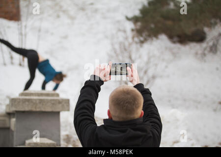 Teenager boy makes photo on smartphone of acrobatic jump girl in winter city park - parkour concept Stock Photo