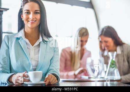 Portrait of beautiful young businesswoman having coffee with colleagues in background at cafe Stock Photo
