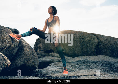 Fit african woman standing by rock at the beach after workout session. fitness woman training outdoors on rocks. Stock Photo