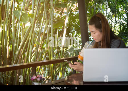 Woman hold mobile phone and drinking iced tea in coffee shop, laptop on wooden table of brown Stock Photo