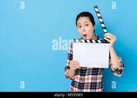 beautiful young woman playing clapboard standing in blue background and looking at camera making surprised emoticon face. Stock Photo