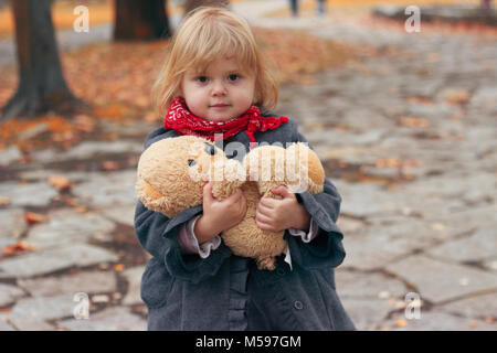 Beautiful little girl holding her teddy bear in park, on cold autumn day; autumn background Stock Photo