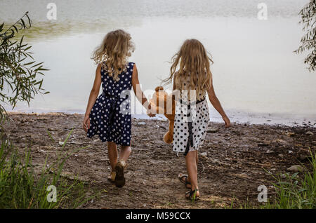 two little girls with long blond hair holding a teddy bear toy and running to the water Stock Photo