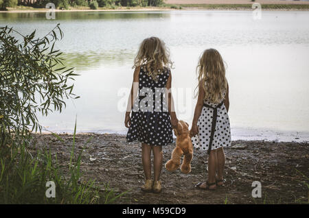 two little girls with long blond hair holding a teddy bear toy and running to the water Stock Photo