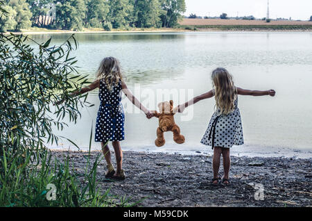 two little girls with long blond hair holding a teddy bear toy and dancing Stock Photo