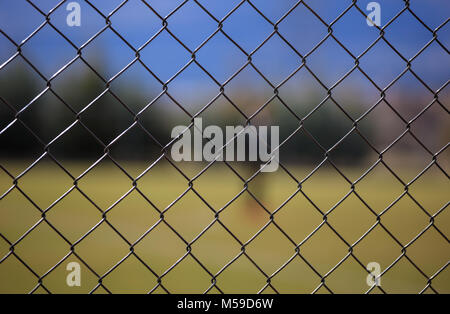 Wire metallic mesh fence around a football field. Blurred green grass, blue sky and trees background, close up view with details. Stock Photo