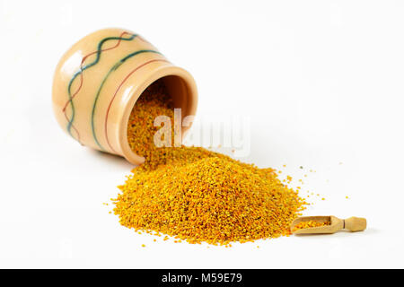 Heap of bee pollen granules, a wooden scoop and an overturned glazed clay pot full of pollen. Selective focus. Closeup. Stock Photo