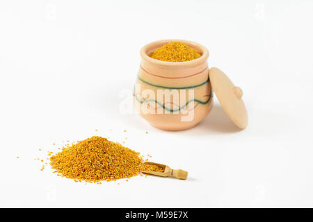 Pile of bee pollen and a little wooden scoop. In a blurry background, it is visible glazed clay pot full of pollen with a lid leaning against the pot. Stock Photo