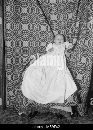 Infant in a christening gown portrait, ca. 1905. Baby is seated on hidden mother’s lap behind blanket backdrop. Stock Photo