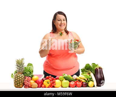 Overweight woman eating a salad behind a table with fruit and vegetables isolated on white background Stock Photo
