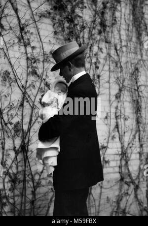 A father tightly holds his newborn  against a backdrop of vines on a wall, ca. 1940. Stock Photo