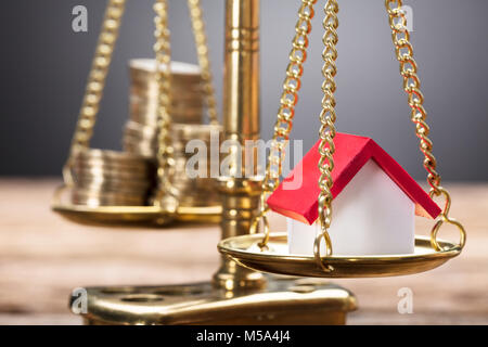 Closeup of model home and coins on golden weighing scale against gray background Stock Photo