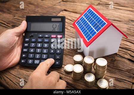 Businessman using calculator by coins and model home with solar panel on wooden table Stock Photo