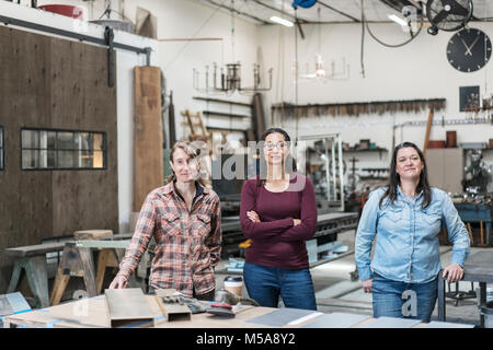 Three women standing in metal workshop, holding looking at camera. Stock Photo
