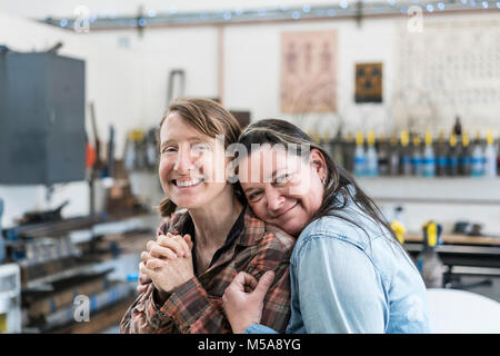 Two women standing in metal workshop, hugging and smiling at camera. Stock Photo
