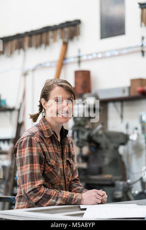 Blond woman wearing checkered shirt standing in metal workshop, smiling at camera. Stock Photo
