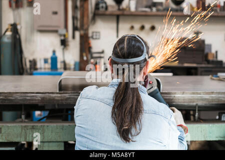 Rear view of woman wearing safety glasses and dust mask standing in metal workshop, using power grinder, sparks flying. Stock Photo