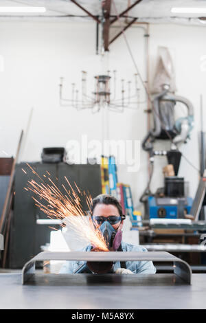 Woman wearing safety glasses and dust mask standing in metal workshop, using power grinder, sparks flying. Stock Photo