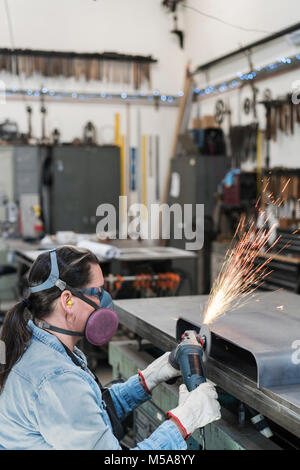 Woman wearing safety glasses and dust mask standing in metal workshop, using power grinder, sparks flying. Stock Photo