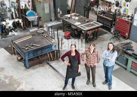 High angle view of three women standing in metal workshop, looking at camera. Stock Photo