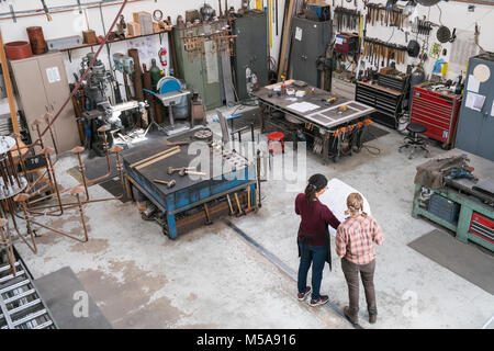 High angle view of two women standing in metal workshop, holding technical blueprint. Stock Photo