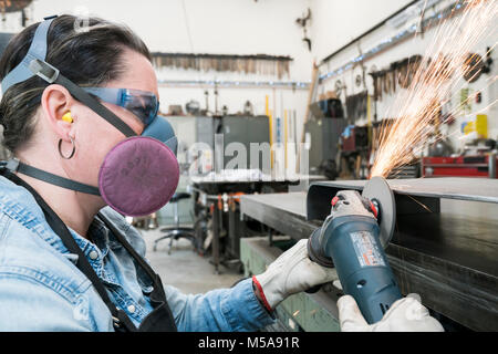 Woman wearing safety glasses and dust mask standing in metal workshop, using power grinder, sparks flying. Stock Photo