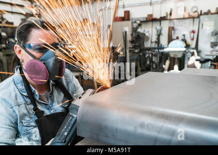 Woman wearing safety glasses and dust mask standing in metal workshop, using power grinder, sparks flying. Stock Photo