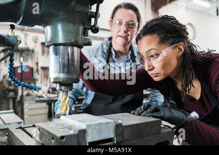 Two women wearing safety glasses standing in a metal workshop, working on metal drilling machine. Stock Photo