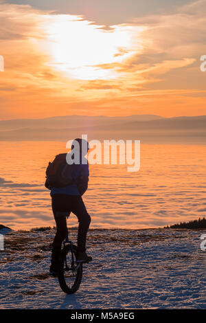 Man on unicycle riding on snowy mountain Schoeckl in Styria, Austria over low stratus fog to sunset Stock Photo