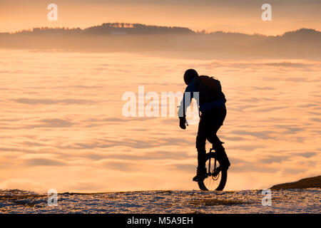 Man on unicycle riding on snowy mountain Schoeckl in Styria, Austria over low stratus fog to sunset Stock Photo