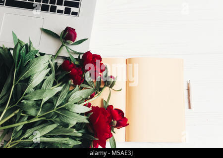 stylish laptop with note book and beautiful red peonies on white wooden background flat lay. space for text. top view. happy womens day or mothers. mo Stock Photo