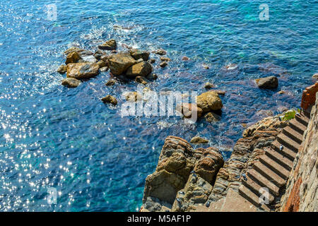 The rocky coastline of Cinque Terre Italy with the sun reflecting off the aqua blue shallow sea while pigeons play on the steps and large rocks Stock Photo