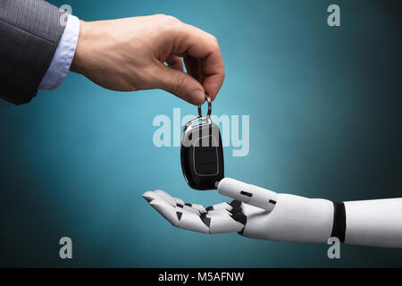 Close-up Of A Businessperson's Hand Giving Car Key To Robot On Grey Background Stock Photo