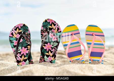 closeup of two different pairs of colorful flip-flops, one of them flower-patterned and the other striped, stuck on the sand of a quiet beach, with so Stock Photo