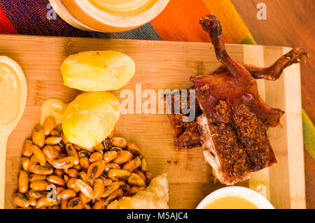 Cut pieces cooked guine pig meat lying on wooden surface next to potatoes, tostados and bowl of salsa Stock Photo