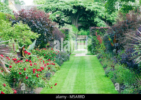 Grass path between flowerbeds with red roses and flowering plants, herbs leading to a metal gate and wooden bench under branchy tree, in a summer Engl Stock Photo