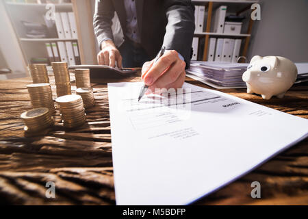 Close-up Of A Businessperson's Hand Signing Document With Stacked Golden Coins On Wooden Desk Stock Photo