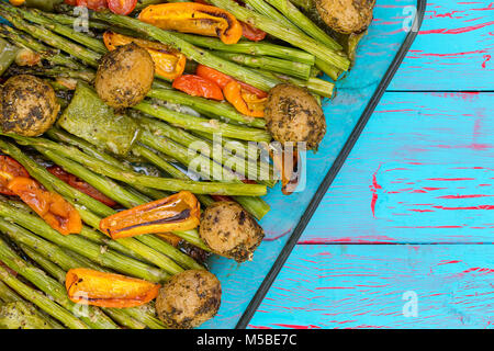 Healthy seasoned roasted fresh vegetables in a glass dish with green asparagus spears, mini bell peppers, tomato and potato over an exotic blue crackl Stock Photo